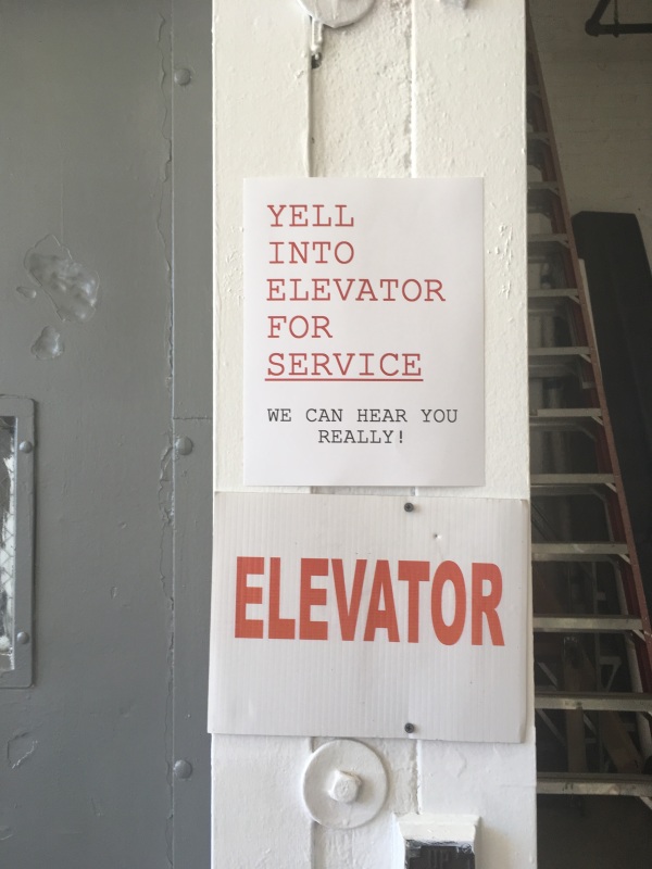Image of two signs on a white wall. One says "Elevator" and the other says "Yell into elevator for service. We can hear you really!"